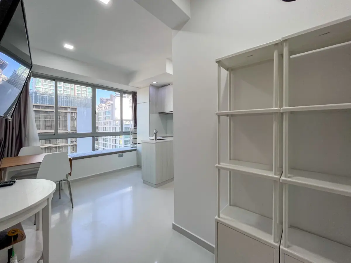Bright, minimalist studio with a large window showing city buildings, a white round table and chair by the window, and a tall open white shelving unit on the right.