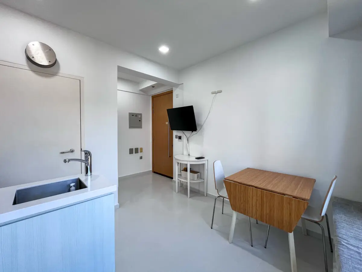 Minimalist studio interior with sink at left, wooden dining table and two chairs, wall-mounted TV near the entrance.