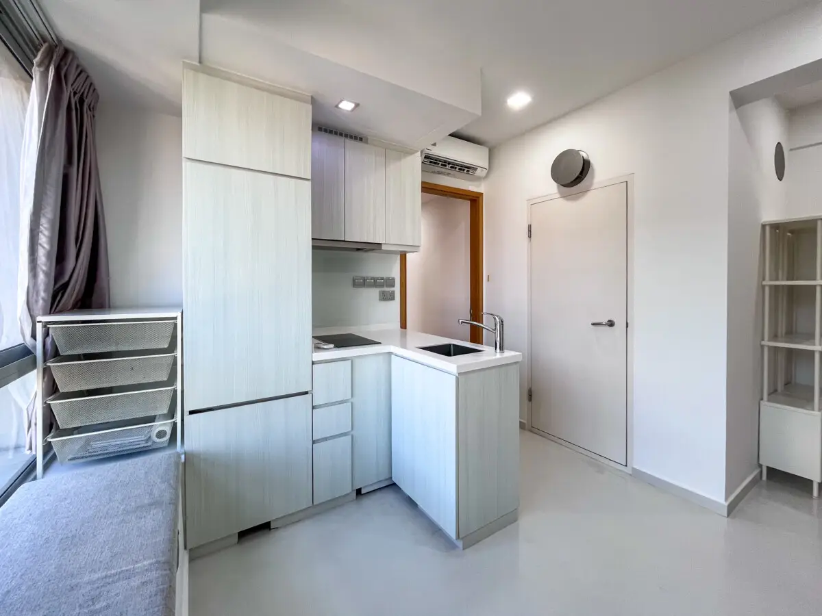 Modern compact kitchen with pale wood cabinets, white countertops, and an island sink area; doorway to the right and a window with curtains on the left.