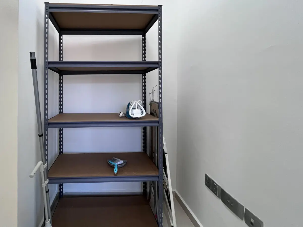 Narrow utility closet with a tall gray metal shelving unit; an iron sits on the middle shelf, a blue scrub brush on the bottom, and a crutch leaning against the left wall.