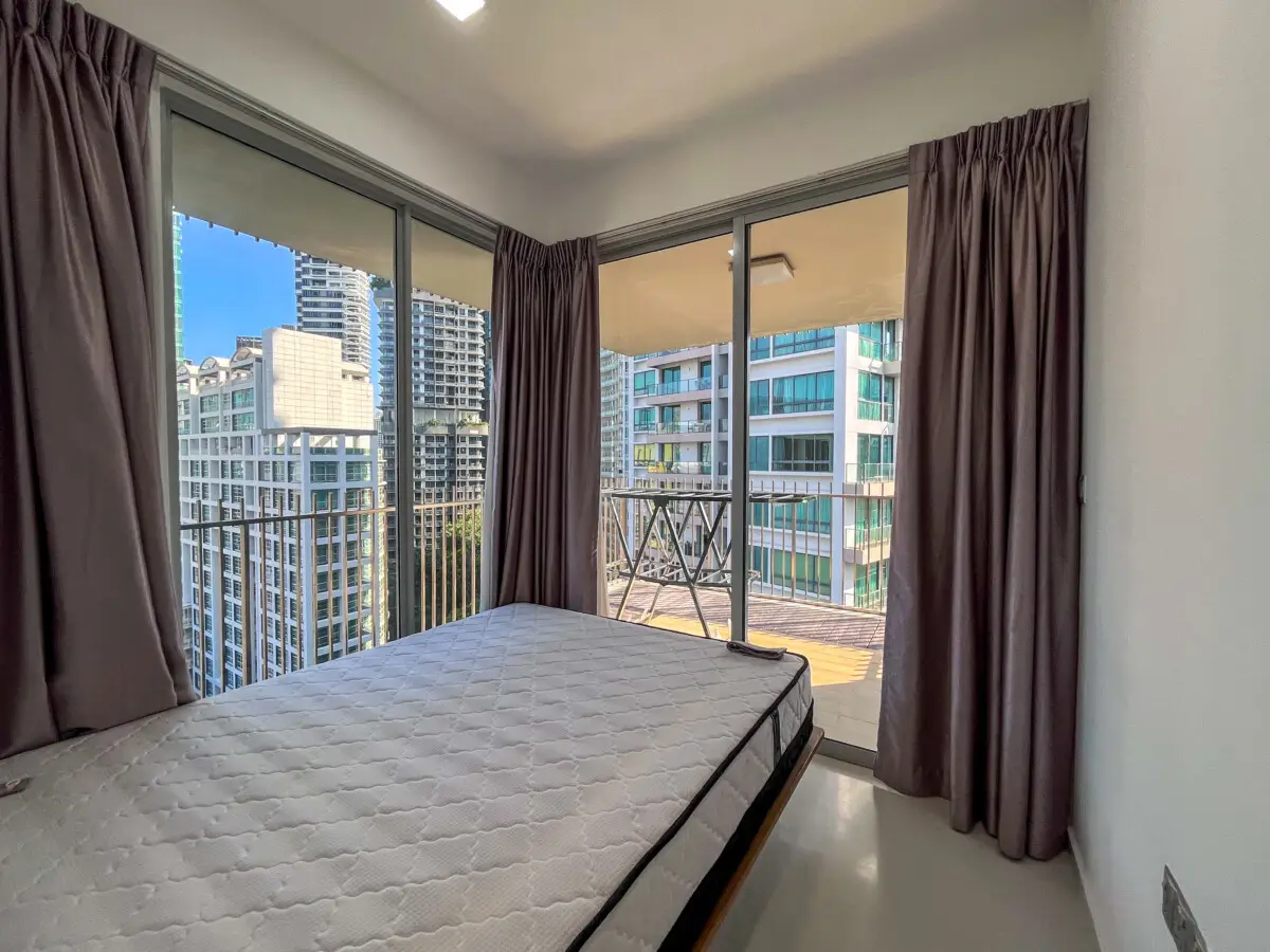 Bedroom with a mattress on a low bed frame, large windows, and a balcony overlooking city high-rises outside.