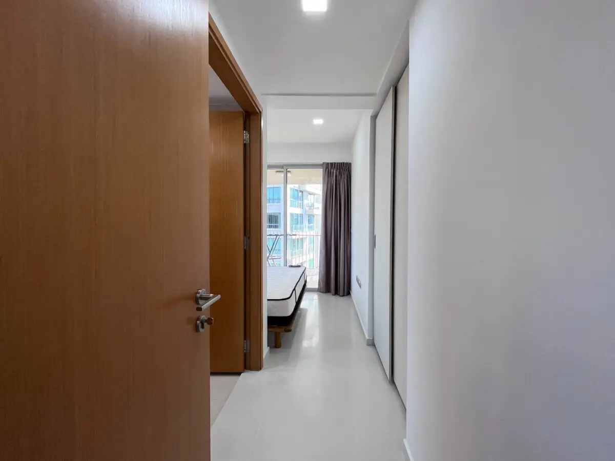 Narrow hallway in a modern apartment leading to a bedroom with a bed by a large window and purple curtains.
