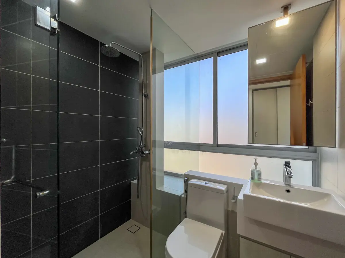Modern bathroom featuring a glass-walled shower, dark tile wall, a white toilet, and a white sink by a large frosted window.