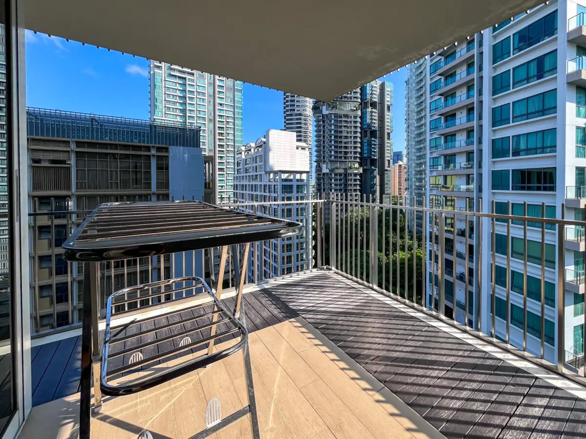 Balcony with a metal drying rack and a metal chair, overlooking tall city buildings on a sunny day.
