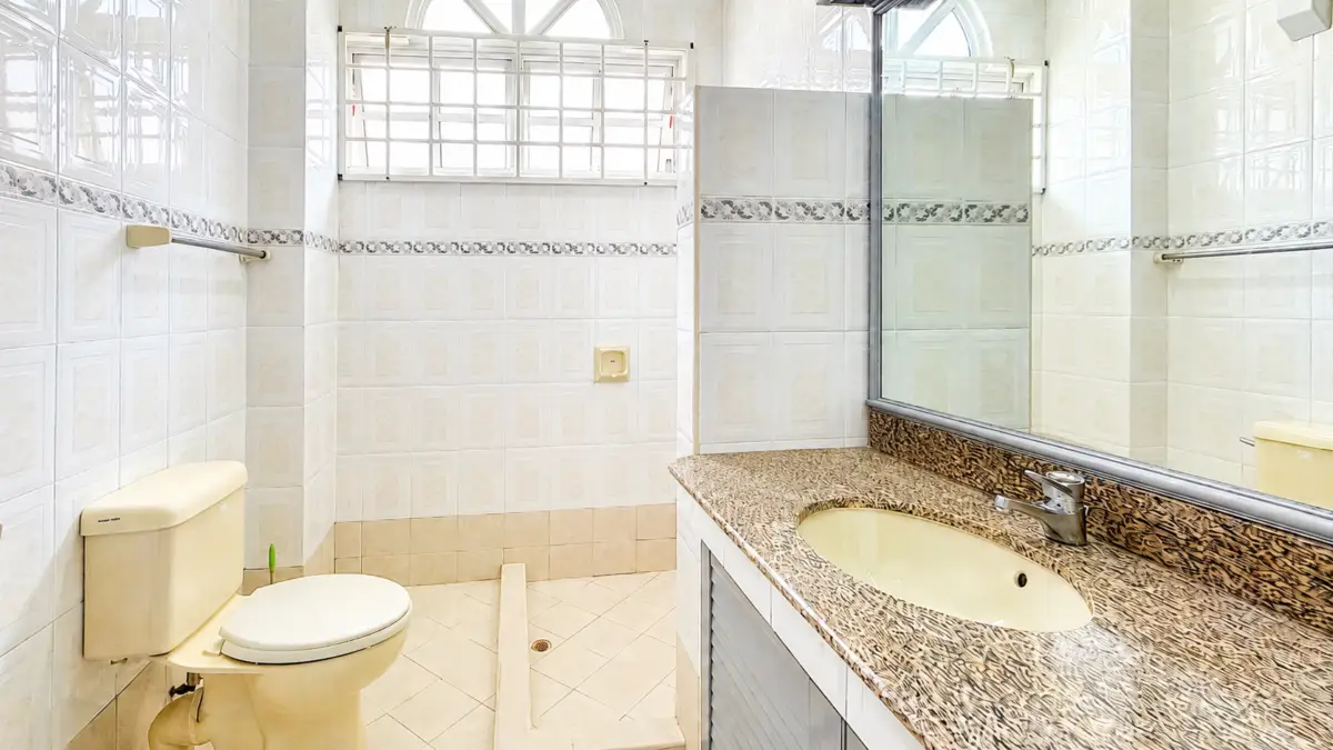 Bathroom with beige toilet on the left and a speckled granite countertop sink with large mirror on the right, all tiled walls and a small window above.