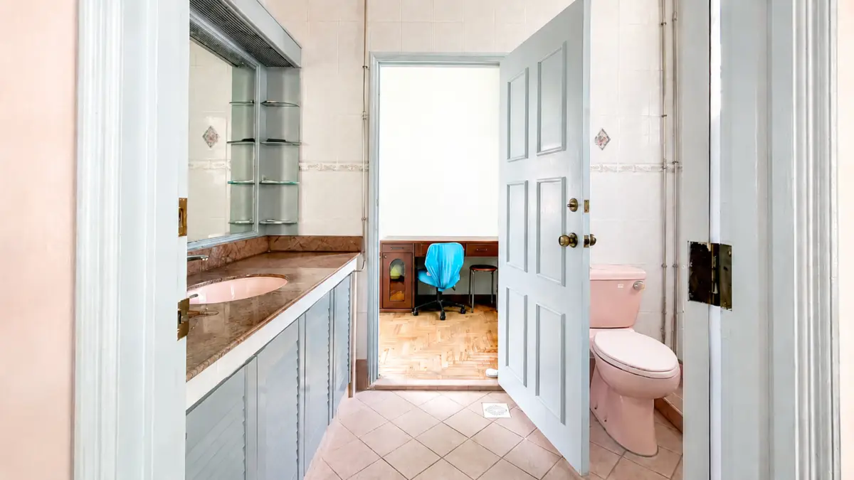 Bathroom with pink toilet and sink, open door to an office area with a blue chair and desk in the next room