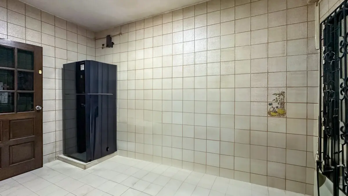 Utility room corner with a tall dark navy cabinet beside a wooden door, all walls and floor tiled in beige. A small decorative tile with a plant is on the right wall, and a metal gate is visible at the far right.