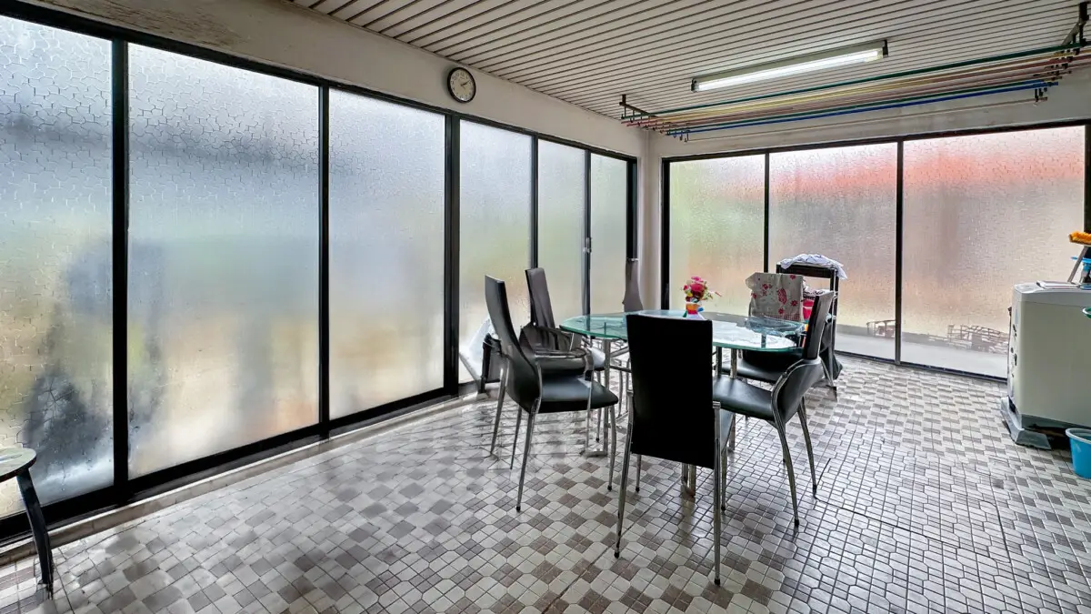 Sunroom with frosted glass walls, round glass table and six black chairs; clock on the wall and clothesline rods overhead.