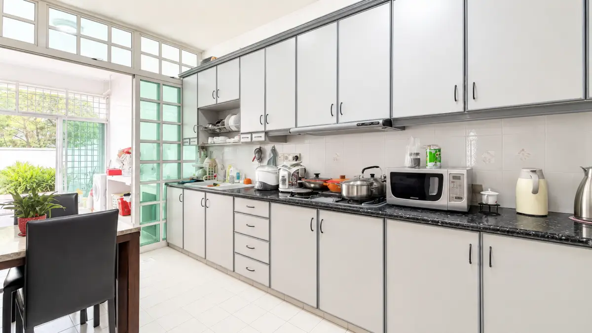 Long kitchen with gray cabinets, black granite counter, and a row of appliances (microwave, kettle) with pots on the stove; a dining table and black chairs sit at left near a sunny glass door.