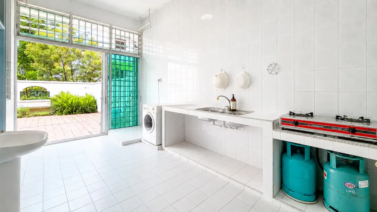 Bright white utility kitchen with a washing machine, sink, and a two-burner gas stove, plus blue LPG cylinders stored beneath the stove. Outdoor patio with greenery is visible through the door.