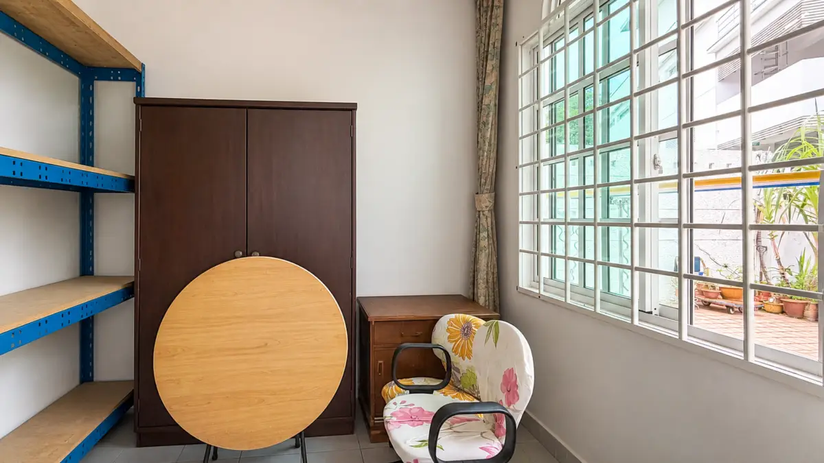 A storage room with blue metal shelves, a dark wooden cabinet, a round wooden table leaning against it, and a floral chair by a large window.