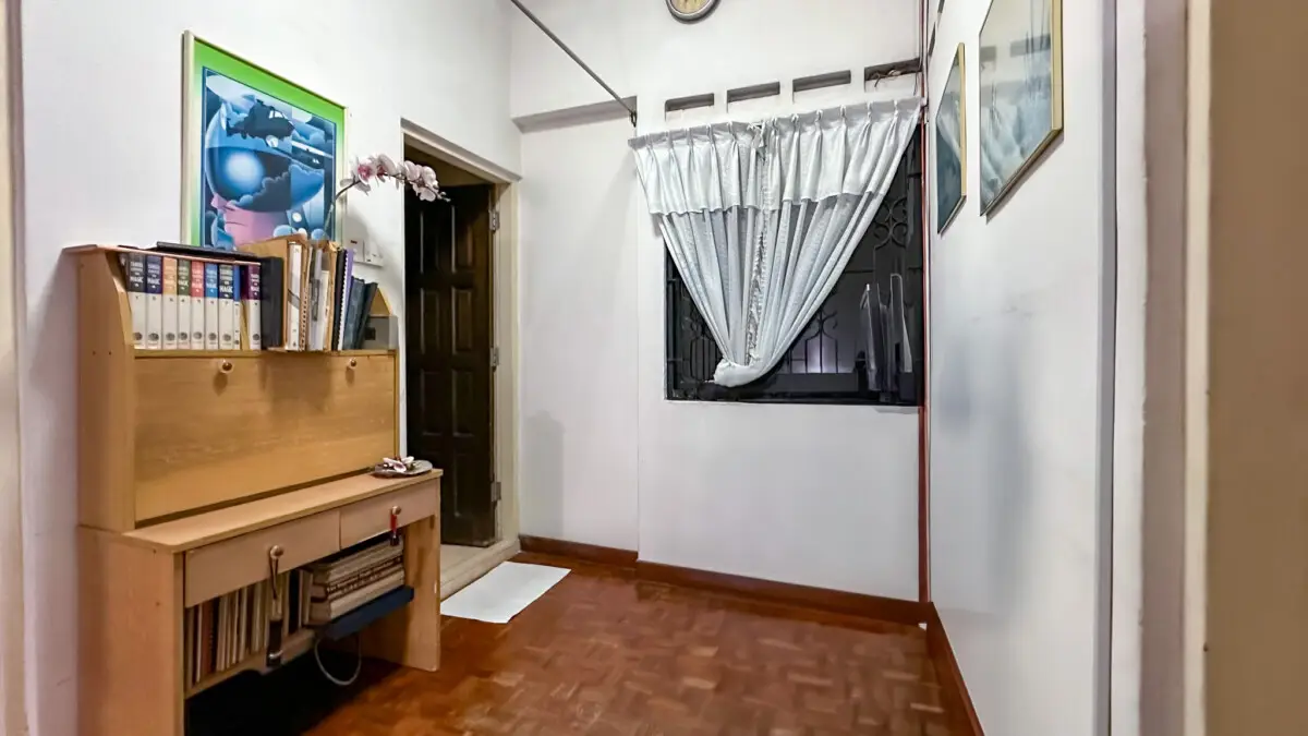 Narrow entry hall/study with a wooden desk on the left, binders, and books; dark door, window with white curtain in the back; framed pictures on the right wall.