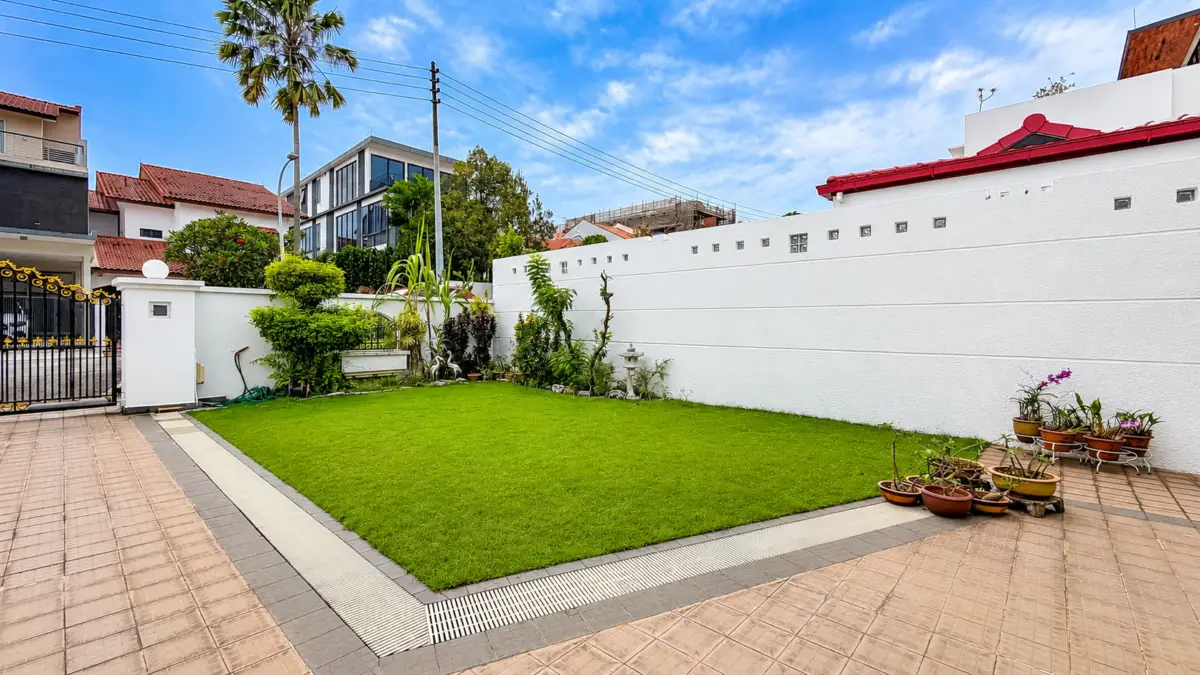 Front yard with a rectangular patch of green grass, potted plants along a white boundary wall, and tiled paving on the sides.