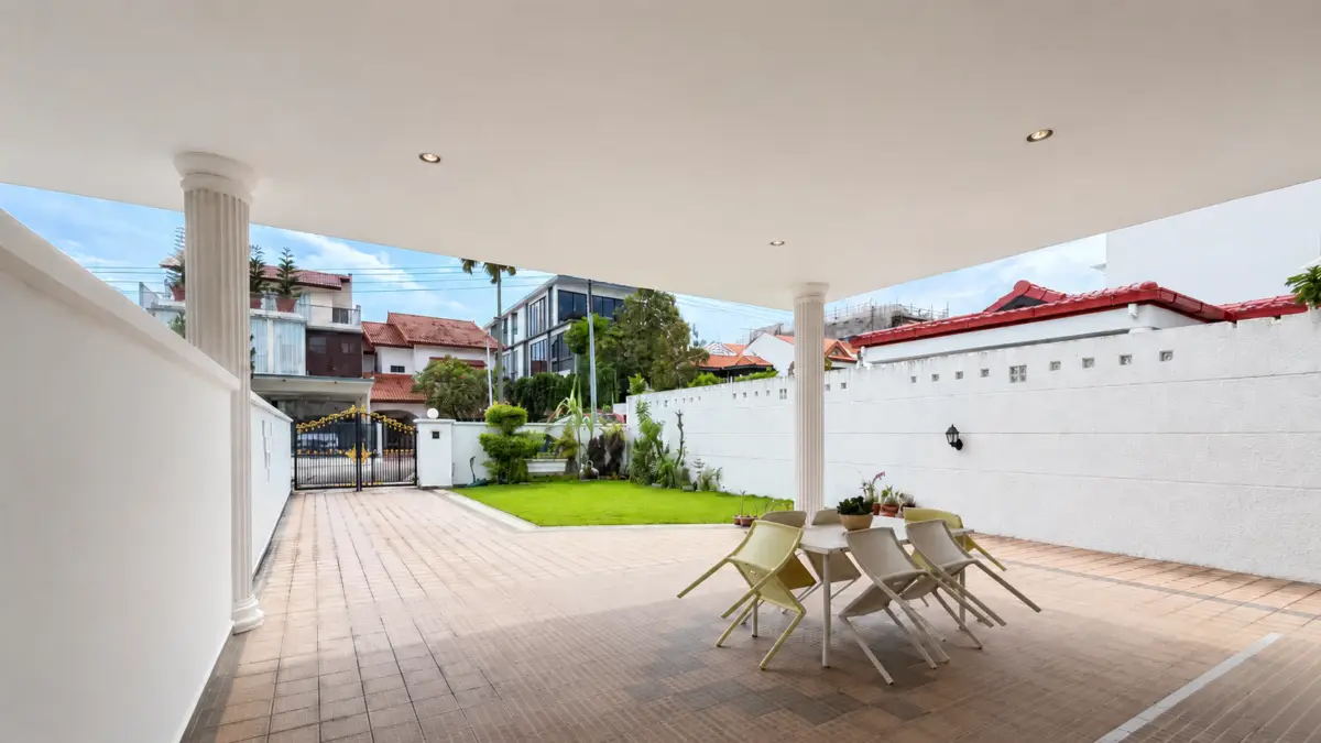 Covered outdoor patio with round dining table and tan chairs on a tiled floor, white walls, and a green lawn beyond.