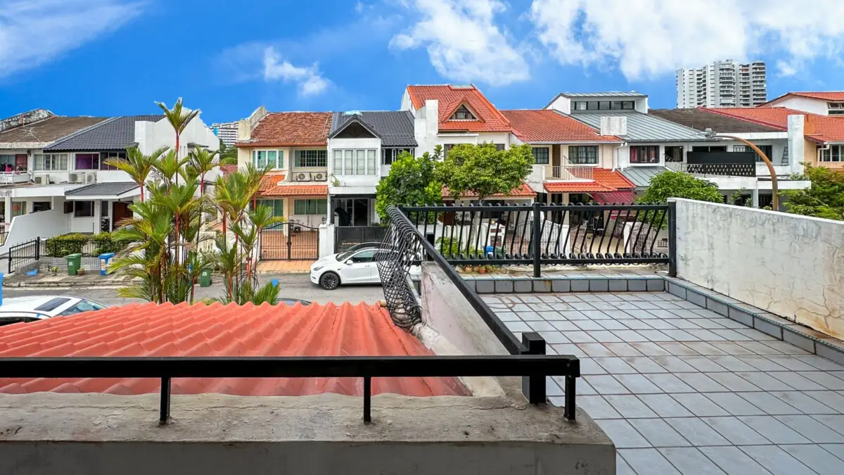 Rooftop tiled terrace with a black railing overlooks a street of colorful two-story houses under a blue sky; palm plants are in the foreground.