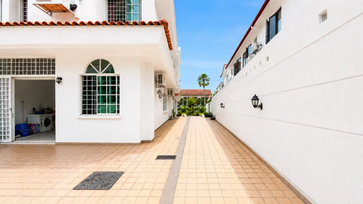 Open-air courtyard between white apartment buildings with tan tile pathway, wall lanterns, and a distant palm tree under a blue sky.