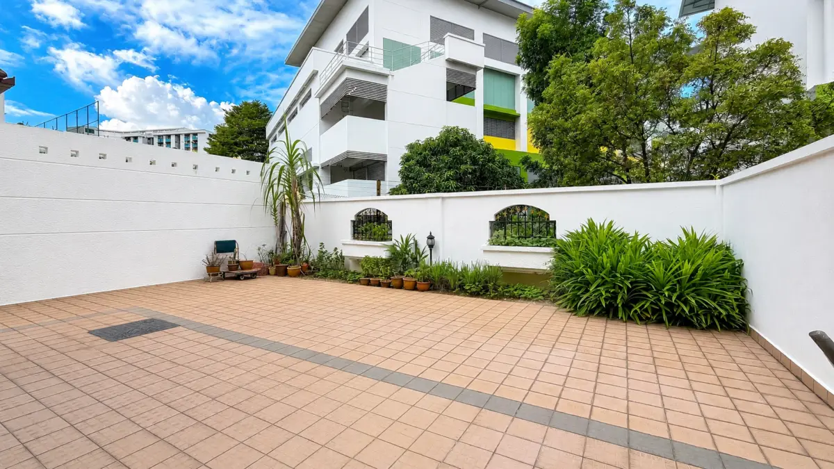 Sunny courtyard with tan tiled patio, white boundary walls, and a row of potted plants along a planter wall; a modern white building with balconies rises in the background under a blue sky.