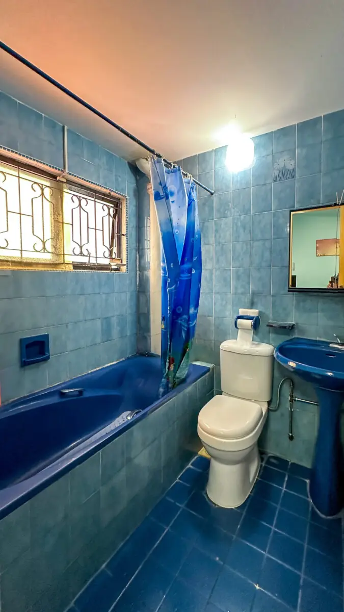 Blue-tiled bathroom with a blue bathtub, toilet, and sink; blue shower curtain partially drawn beside the tub and a barred window on the left.