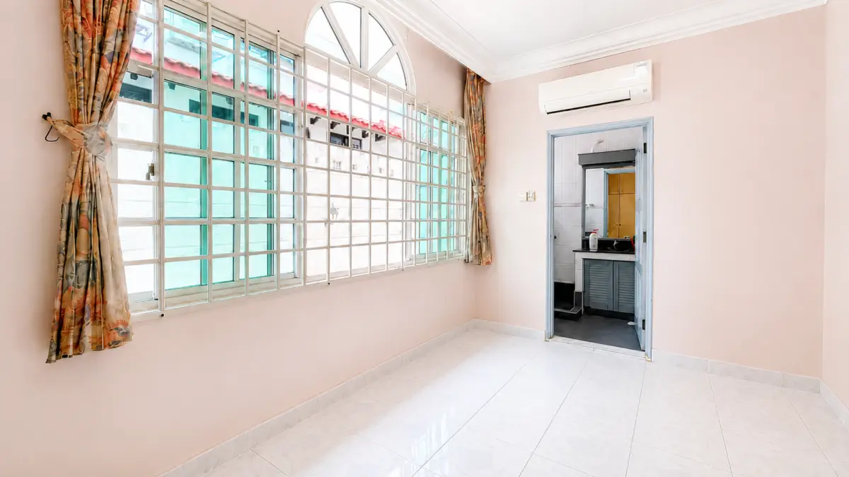 Sunlit empty pink room with a large grid window and floral curtains, white tiled floor, and an open doorway leading to a small kitchen area with a sink and cabinets. An air conditioner sits high on the wall above the doorway.