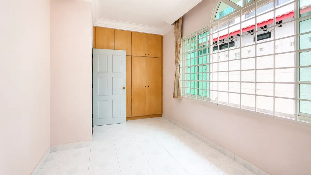 Narrow sunlit room with pastel pink walls, a light blue door, built-in wooden wardrobes, and a large barred window.