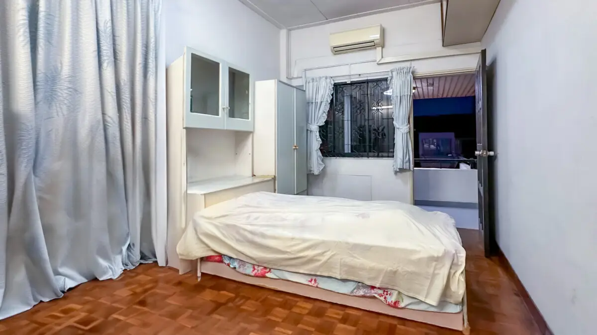 Small bedroom with a single bed, white storage cabinets, and a window with bars; blue curtains and an open door to a balcony at night.