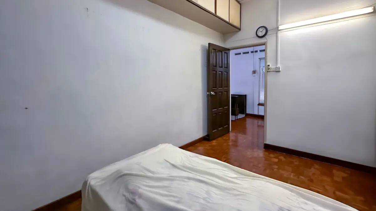 Sparse bedroom with a bed in the foreground, white sheets, and an open door revealing a small desk area beyond.