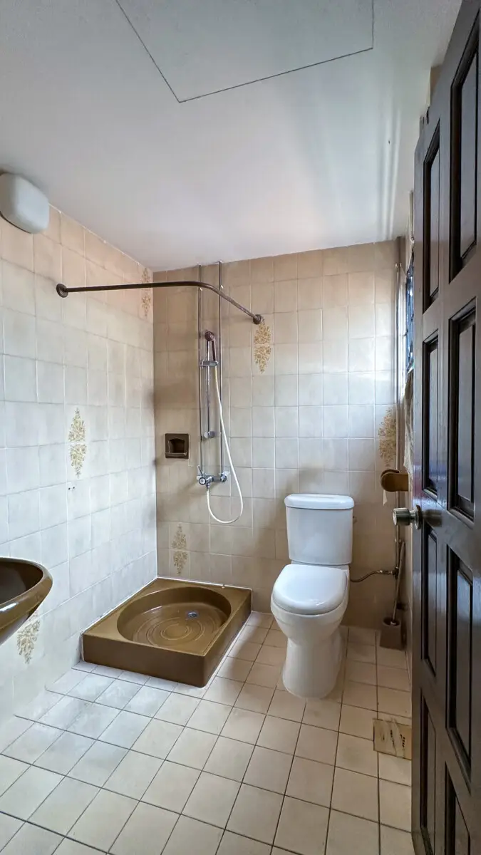 Compact beige-tiled bathroom with a brown raised squat shower tray, wall-mounted shower, white toilet, and sink; dark wooden door on the right