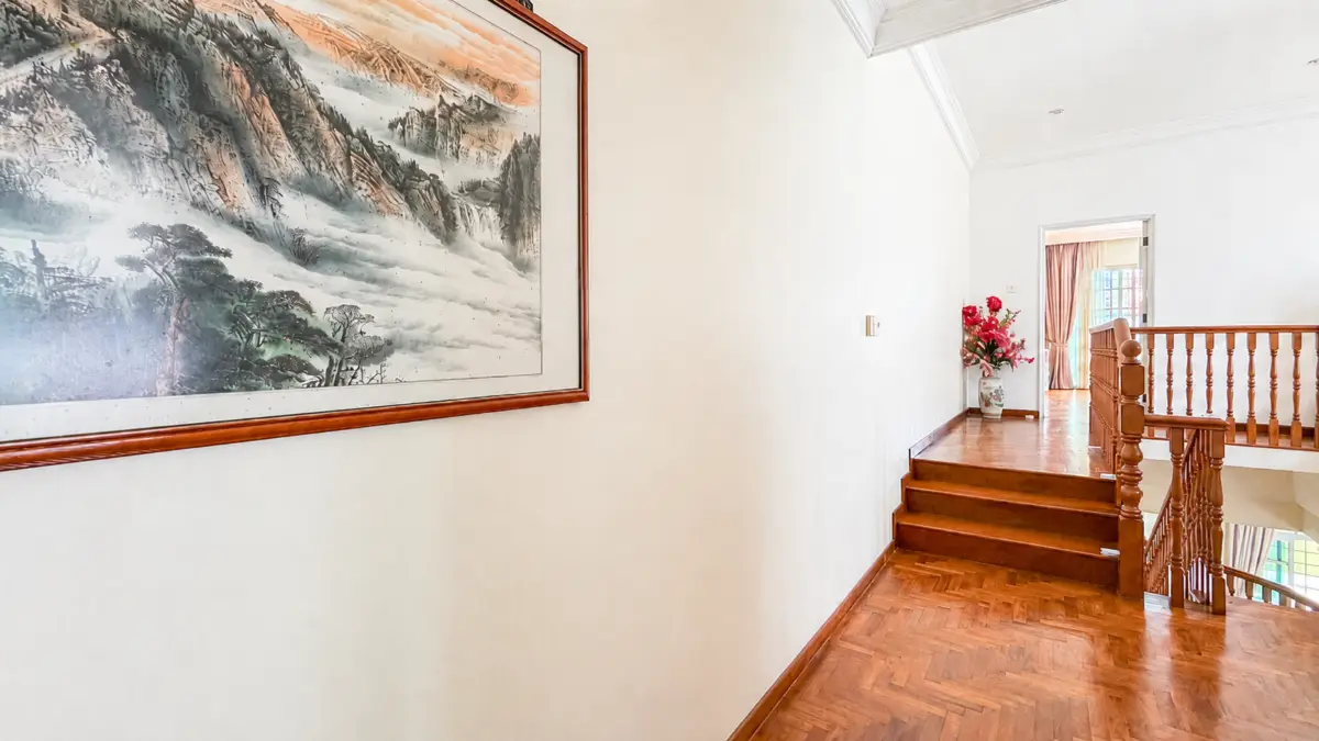 Bright hallway with a large landscape painting on the left, dark wood floor, and a staircase railing at the end with a vase of red flowers nearby.