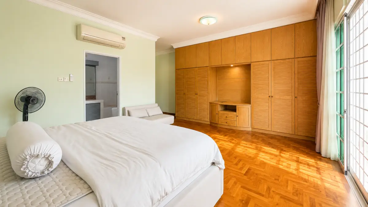 Bright bedroom with a large white bed, a standing fan, pale green walls, and a wall of built-in wooden wardrobes of varying doors…