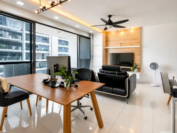 Bright modern home office with a wooden desk, computer, and a potted plant near large windows and a black leather sofa beyond.