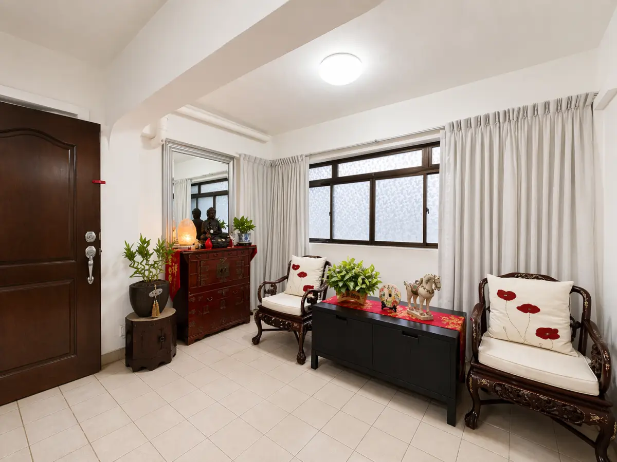 Cozy living area with white walls, a dark wooden door, potted plants, a red carved chest, and two ornate chairs with floral cushions by a black console table.