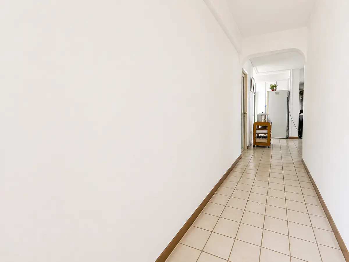Long white hallway with beige tiled floor leads to a small kitchen area with a metal refrigerator and a wooden cart at the end.