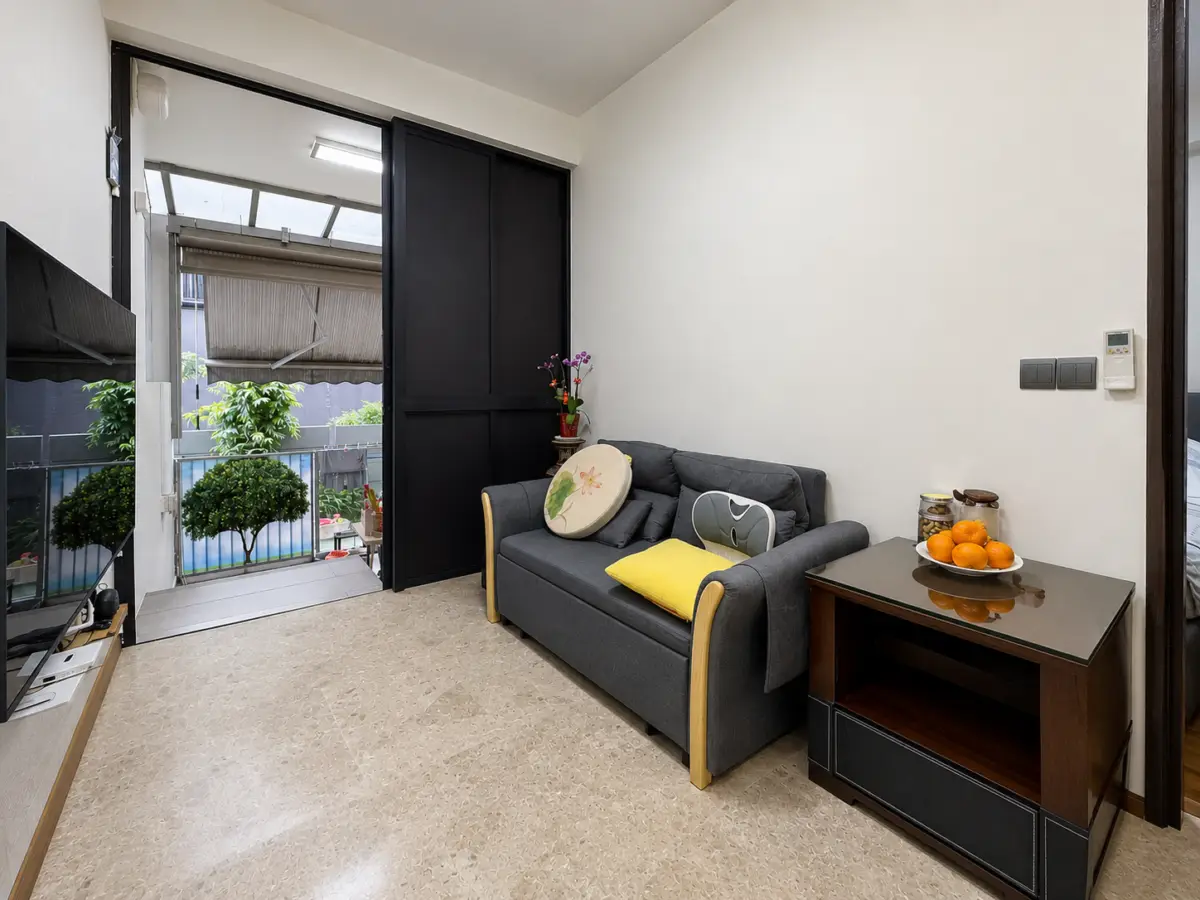 Cozy living room with a dark gray sofa, yellow pillow, and a round decorative cushion; a glass-top side table holds a fruit bowl and jars, with an open balcony door showing potted plants outside.