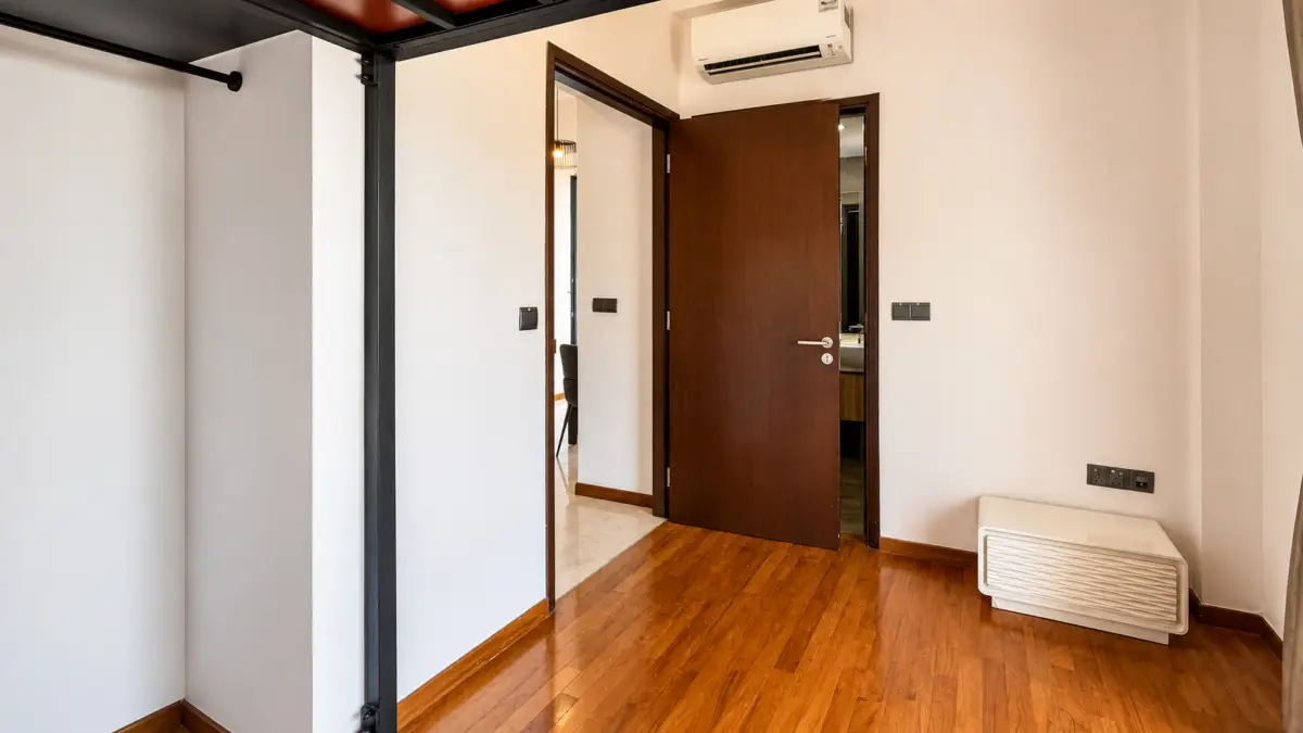 Room interior with a partially open brown door, white walls, and warm wooden flooring; an air conditioner is mounted high on the wall above the door."​,
