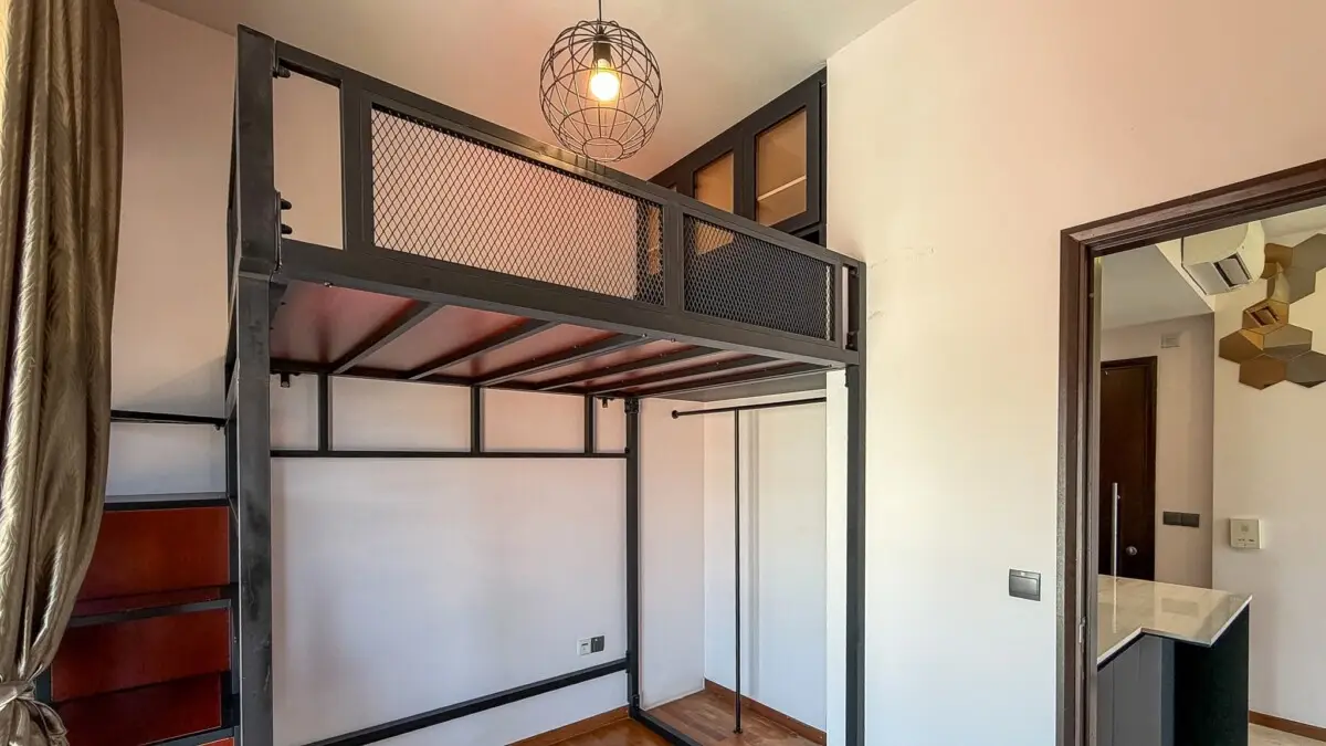 Industrial loft bed in a compact room: black metal frame, upper bunk with mesh guard, and a pendant wirelight overhead.