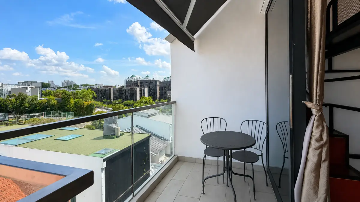 Balcony with a small round metal table and three chairs, overlooking a cityscape under a blue sky with scattered clouds.