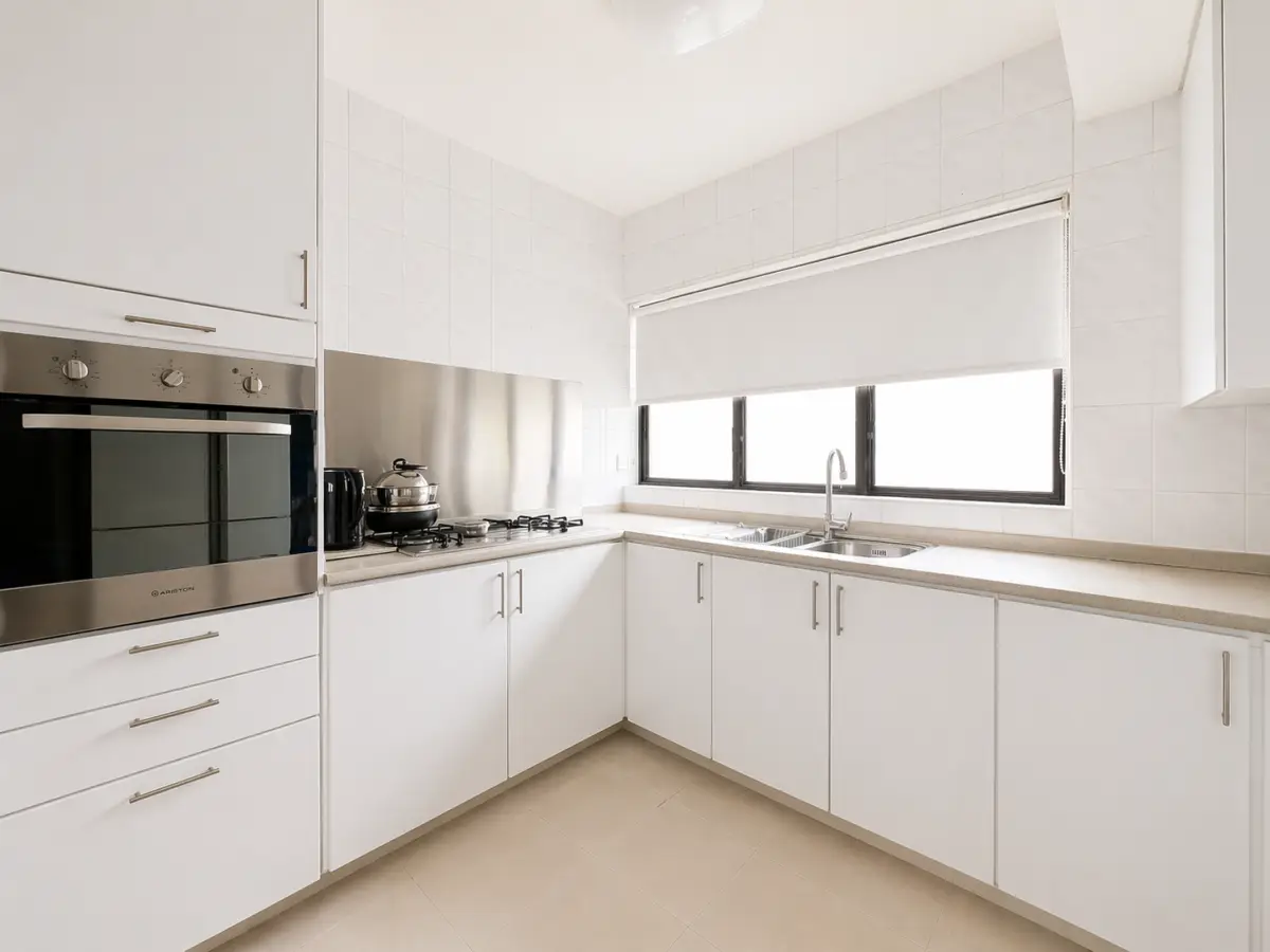 Minimalist white kitchen with built‑in oven, gas hob, and a long beige countertop along white cabinets under a windowed sink area.