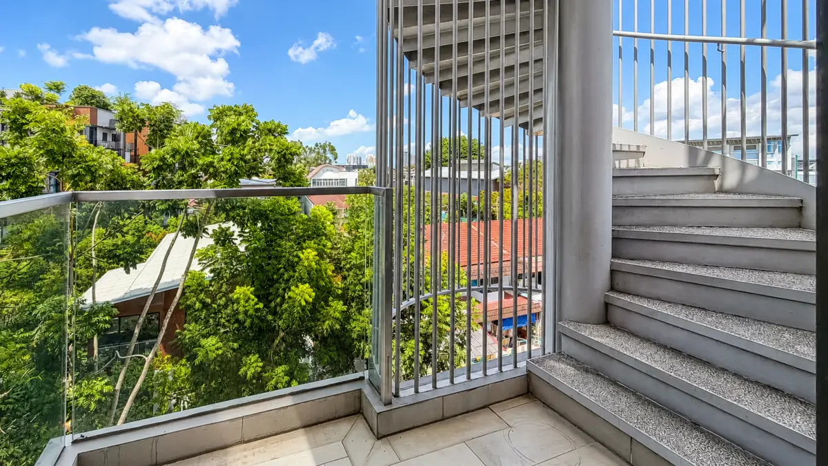 Balcony with glass railing and metal stairs, overlooking green trees and red rooftops under a blue sky.
