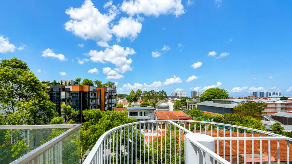 Urban balcony view over rooftops and trees under a bright blue sky with fluffy clouds.