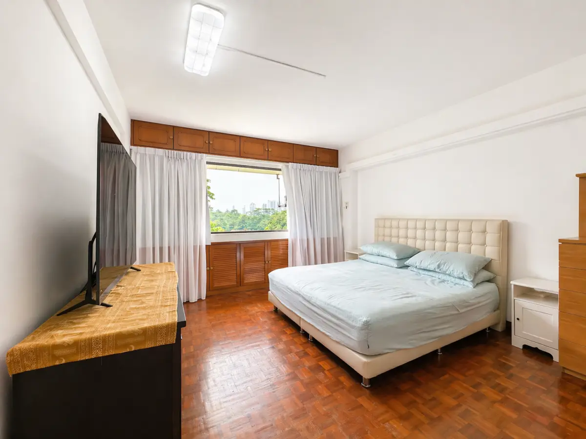 Bright bedroom with a tufted beige headboard bed in pale blue bedding, white nightstands, and a TV on a wooden dresser facing the bed; window with white curtains beside.