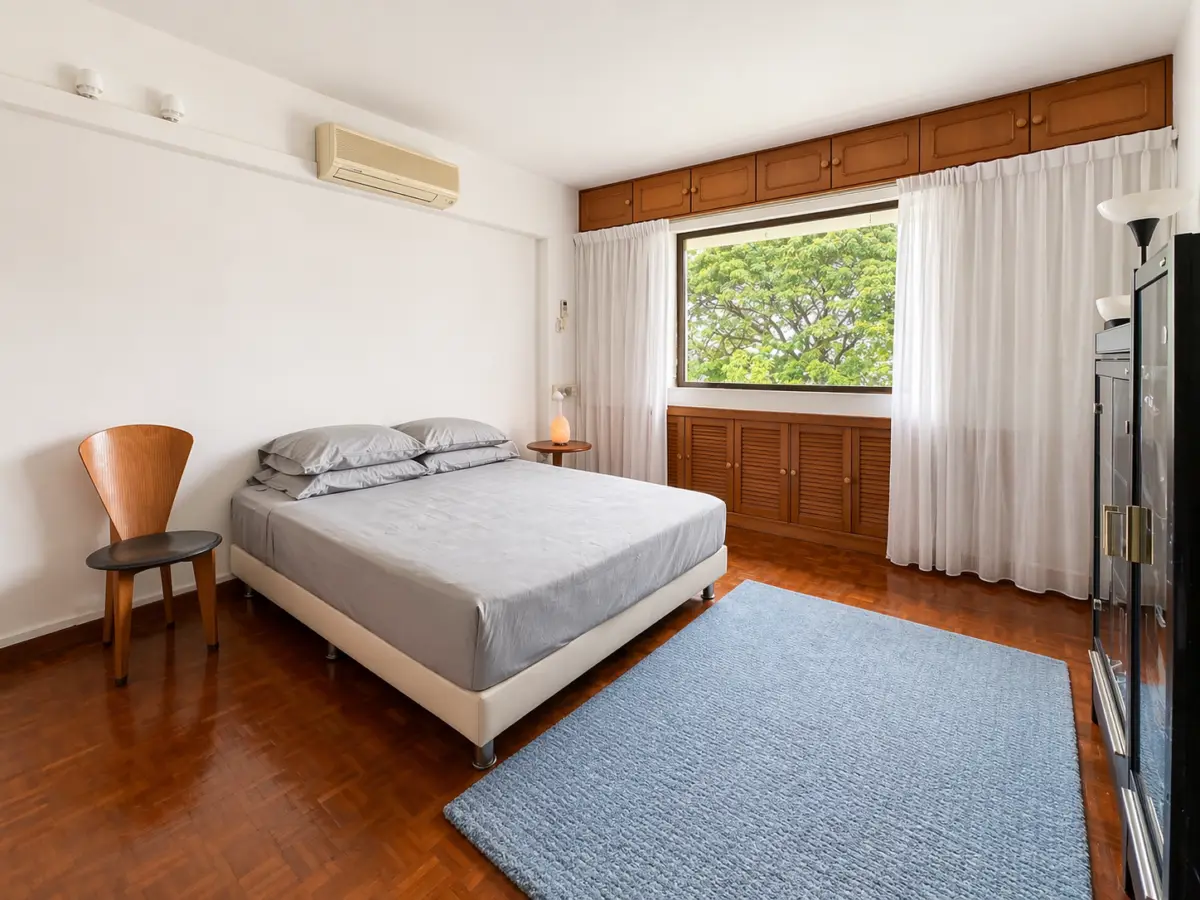 Bright bedroom with a double bed, gray bedding, and a blue rug on a wood floor; window with white curtains and wooden cabinets above and below.