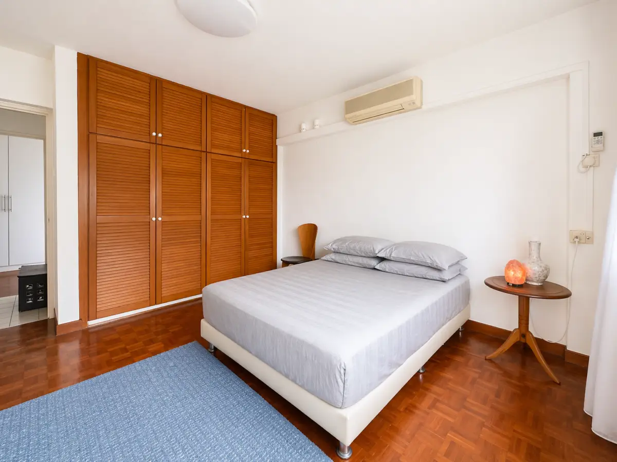 Double bed with gray bedding beside a round wooden bedside table with a lamp, against a white wall and a large wooden wardrobe behind.