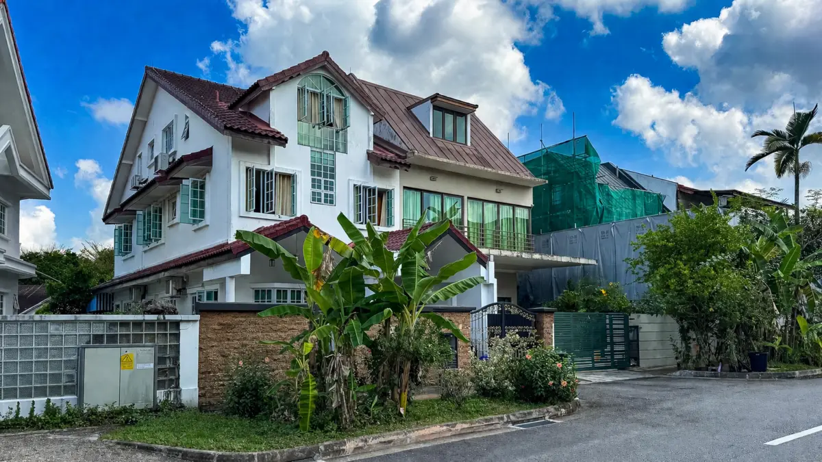 White multi-story house with a red-tiled roof, banana plants in the foreground, and a gated driveway under a bright blue sky.