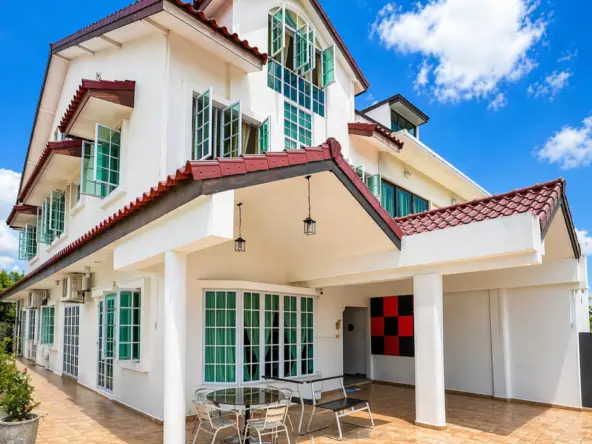 White multi-story house with red-tiled roof and many green-framed windows under a bright blue sky; tiled patio with glass table and chairs divided by a covered entryway.