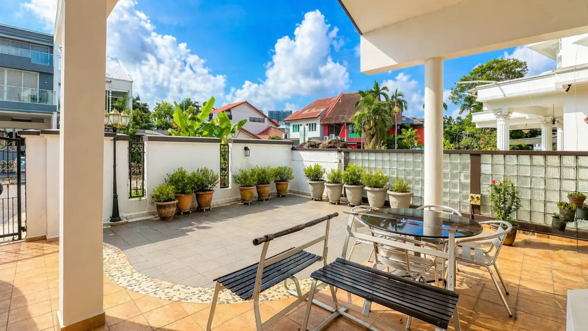 Outdoor patio area with a round glass table and metal chairs under a shaded overhang; row of potted plants along a white wall, clear blue sky above.