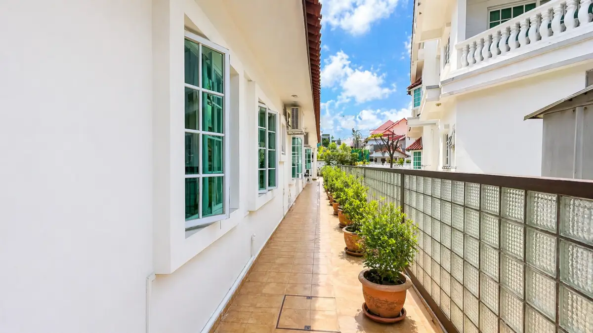 Sunny outdoor corridor with white buildings on the left and potted green shrubs along a tiled path, beside a glass-block wall on the right.