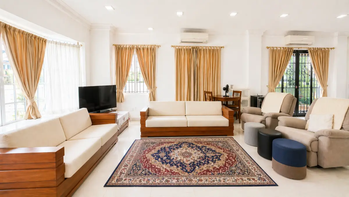 Bright living room with beige sectionals, wooden frames, a patterned area rug, and large windows with tan curtains to let in natural light.