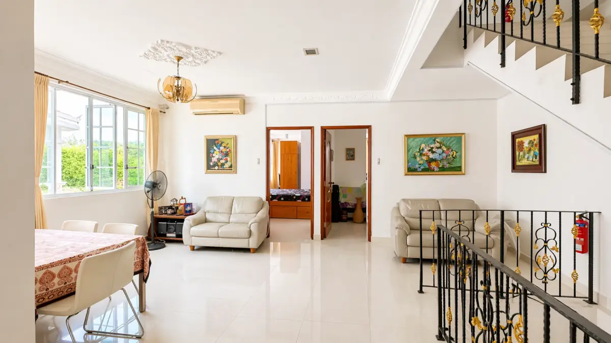 Bright living room with white tiled floor, two beige sofas, a dining table, and a large window with beige curtains. Outside greenery visible.