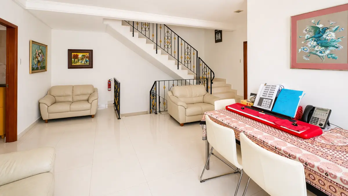 Bright living room with beige sofas, a stairway railing, and a dining table covered by a patterned tablecloth with office items on it.