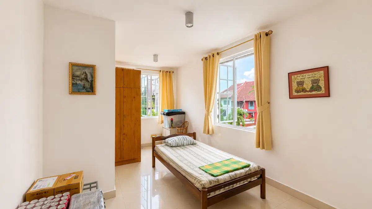 Small bright bedroom with a single wooden bed, striped bedding, and a wooden wardrobe by the wall under two windows with yellow curtains: a tidy, lived-in feel.